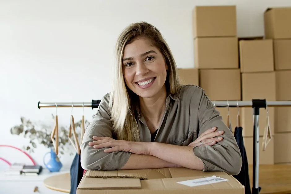 Happy woman managing her online store with packages and clothing rack.